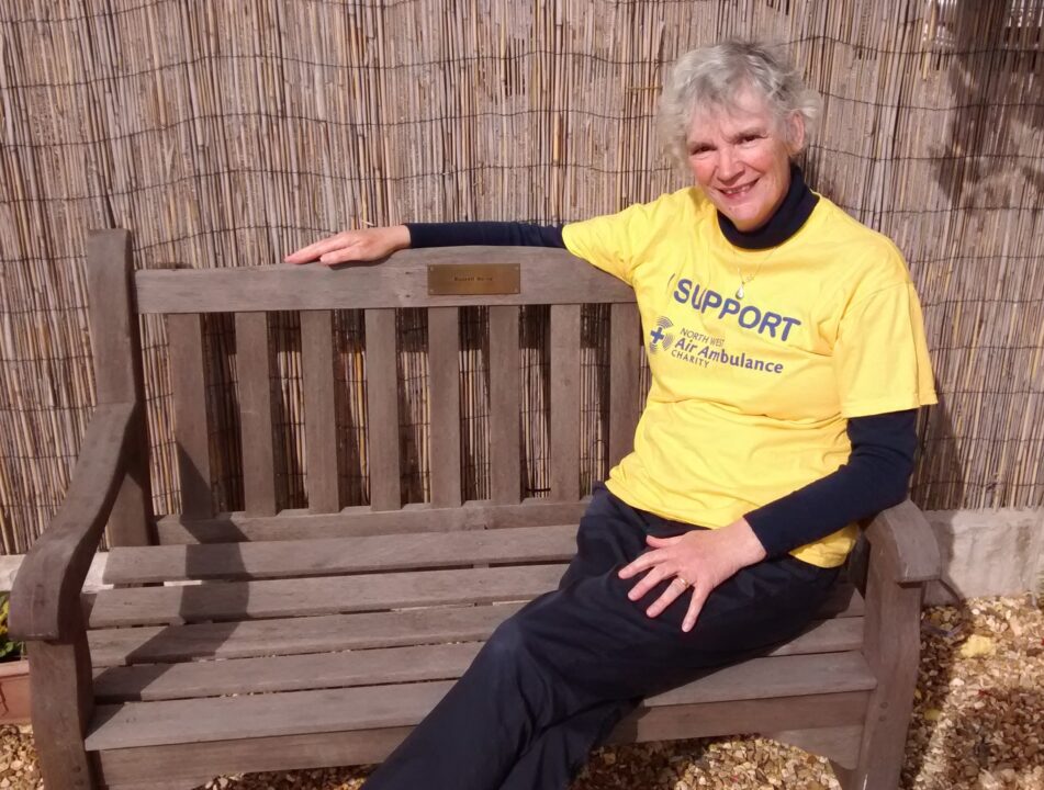 Volunteer Sarah, dressed in a yellow t-shirt sat on a bench dedicated to her Husband 