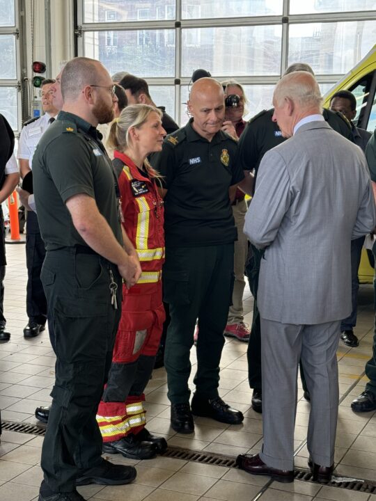 a group of people stood together, two are in dark green outfits, one is in a red suit and the other is in a grey suit - His Majesty meeting emergency responders