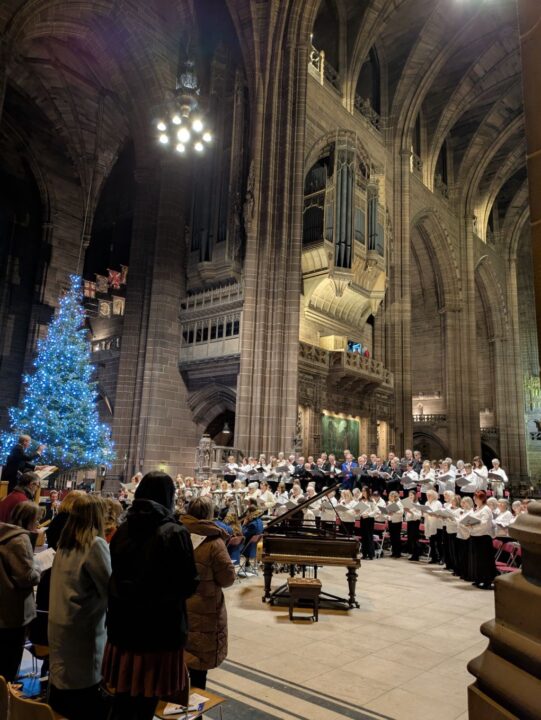 A view from inside Liverpool Cathedral, people seated watching the Choir sing with a lit-up chritsmas tree in the back ground