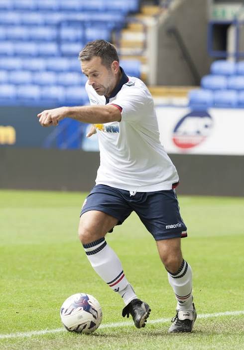 Man dressed in football kit, kicking a football across a football field