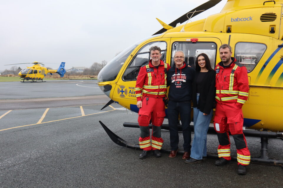 Four people stood in front of the charity's yellow helicopter on a helipad - Two are dressed in red flight suits, and the others are in black and blue jeans