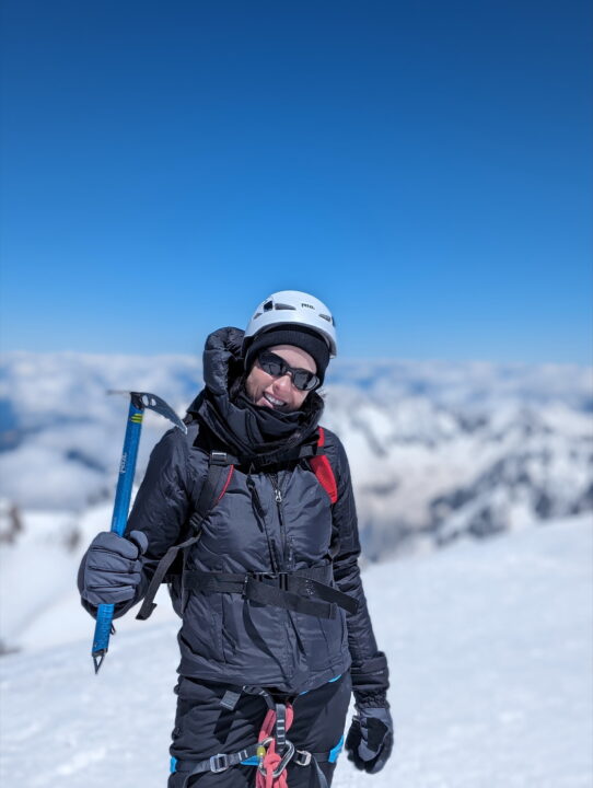 Woman stood on top of a mountain dressed in winter clothing holding a climbing pickaxe 